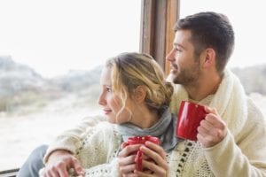 A_happy_couple_with_mugs_and_sweaters_looking_out_at_a_winter_scene_from_their_cabin