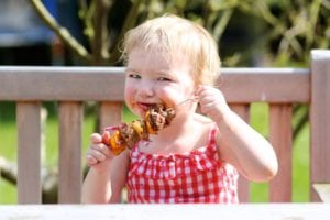 blonde toddler in red gingham top eating barbecue on a skewer