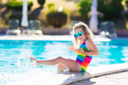Young girl in colorful bathing suit and sunglasses splashing in swimming pool